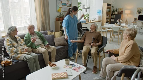 Wide angle shot of female nurse in scrubs talking to seniors in retirement home common room. Suitable for senior care facility and medical insurance advertising