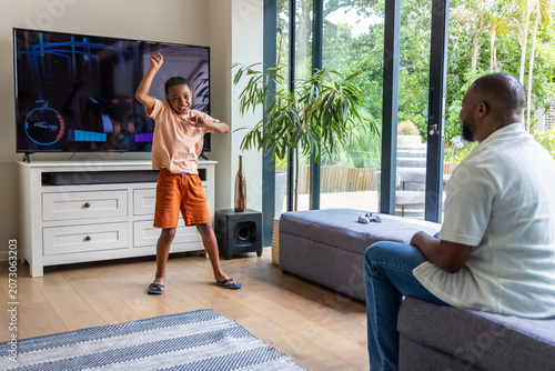 Foto African American father sitting on ottoman watching child dancing near TV at hom