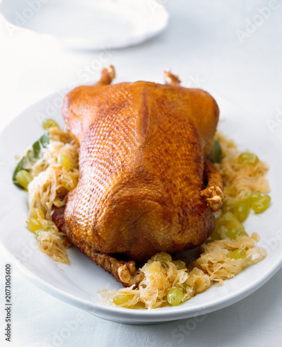A roasted goose rests on a white plate surrounded by pickled cabbage. The dish shows crispy skin and is garnished with green grapes. This meal may be enjoyed at a festive gathering.