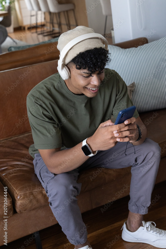 Naklejka premium African American male sitting at home on brown sofa, wearing headphones and holding blue smartphone