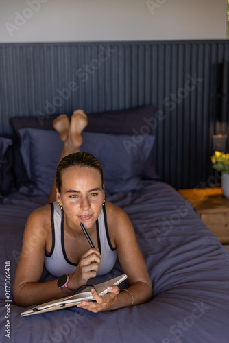 Adult woman lying on stomach on bed in bedroom holding notebook and pen, thinking