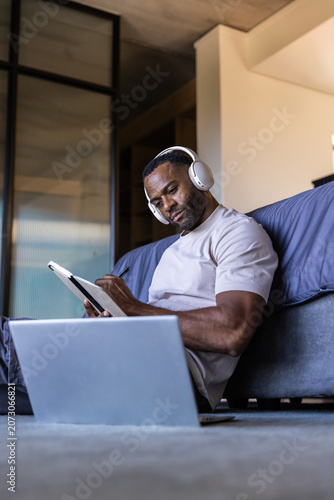 Mature African man wearing headset sitting on bedroom floor using tablet with stylus, open laptop