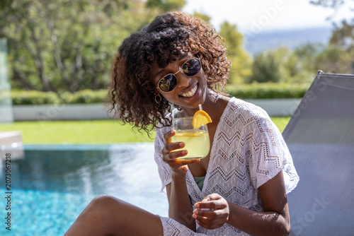 Female sitting poolside wearing shades and white crochet cover-up holding glass with orange slice