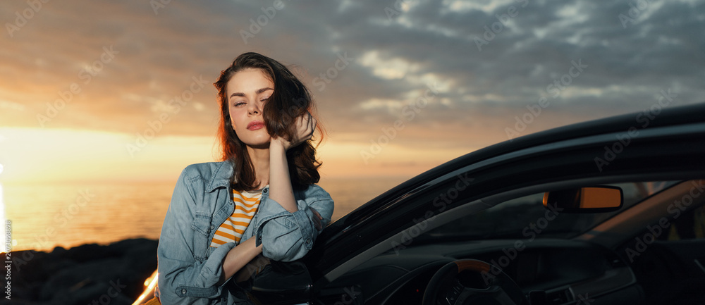 Fototapeta premium Relaxed woman leaning against a car at sunset, denim jacket striped top, wind in hair. A lifestyle moment highlighting travel, freedom, and outdoor evenings.