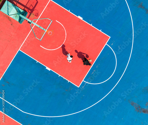 aerial view of two students playing one-on-one basketball on a vibrant red and blue school court. Bright sunlight casts long dramatic shadows, highlighting active outdoor sports and fitness.