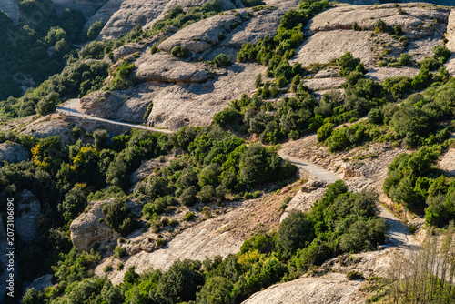 Bild auf Leinwand A high-angle view of a rugged hiking trail winding through the steep, rocky slopes and Mediterranean vegetation of the Montserrat mountain range, Catalonia, Spain