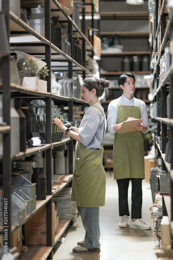 Fototapeta premium A young saleswoman and salesman walk a homeware aisle reviewing stock, the woman noting items on her clipboard while the man scans the bottles, deep among the black shelving