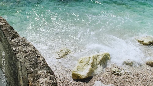 Waves splashing around rocks next to a concrete seawall on a pebble shore, top-down handheld shot, clear turquoise water.