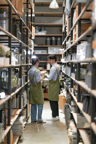 Photography Two young employees in green aprons confer quietly among shelves of pottery and