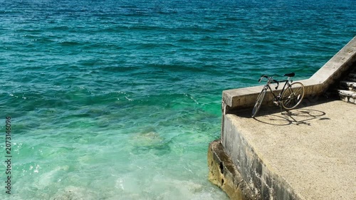 Vintage bicycle parked on a concrete pier edge over clear turquoise sea, casting a shadow on sun-warmed stone, handheld shot.