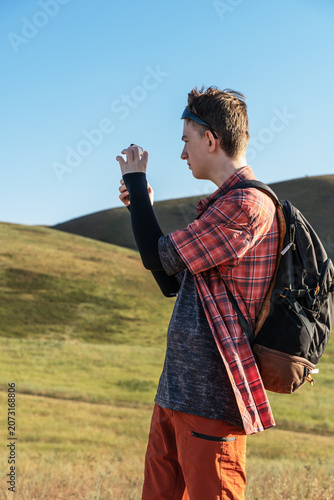 Foto A young teenage boy traveler is standing on a grassy hill in Kyrgyzstan mountains under a clear blue sky, holding a smartphone and taking a picture or filming a video