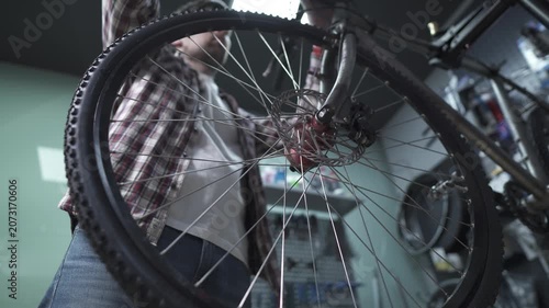 Low angle shot of a male bike mechanic in a plaid shirt repairing and adjusting the front wheel and disc brake system of a mountain bicycle on a special stand inside a professional workshop garage.