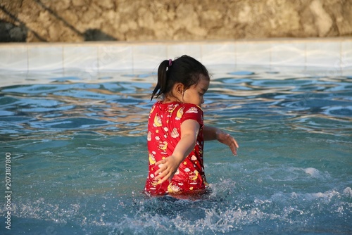 Young girl in a red swimsuit shirt splashes and plays in a clear backyard pool, enjoying warm sunlight and summer fun. Casual candid scene conveying joy, playfulness, childhood and leisure.