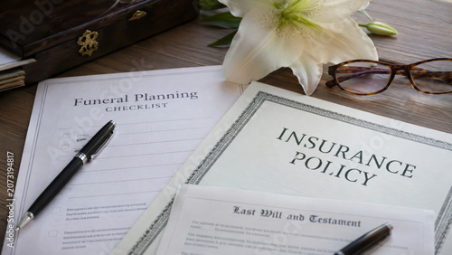 Funeral planning documents with insurance policy, lily and pen on wooden desk. End of life preparation and memorial service paperwork concept.