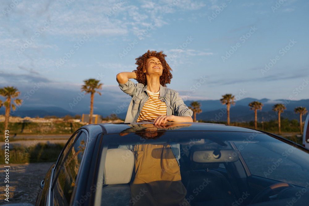 Fototapeta premium Woman leans on car roof under sunny sky, casual denim striped top, open air mood, outdoor lifestyle moment, relaxed and cheerful, bright day, breeze, horizon, nature, palm trees background, candid