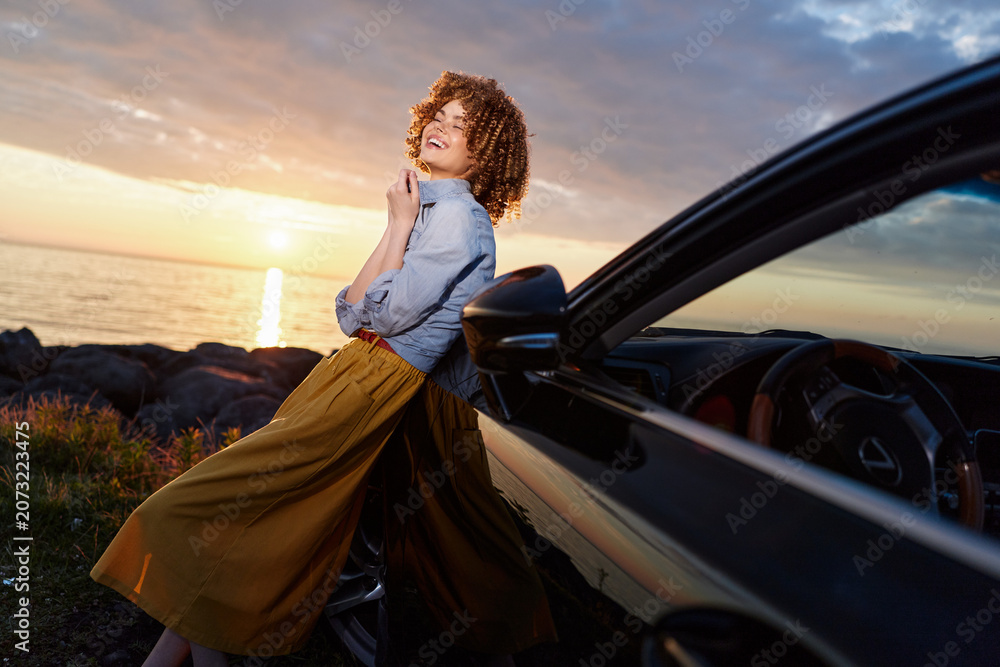 Fototapeta premium Cheerful woman enjoying outdoor drive beside a sleek car at sunset, leaning against the door. Lifestyle moment emphasizing fashion, confidence, and relaxed happiness.
