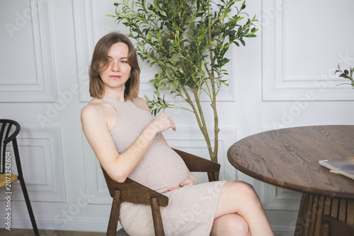 Pregnant woman in beige dress sitting on wooden chair, hold belly in stylish room.