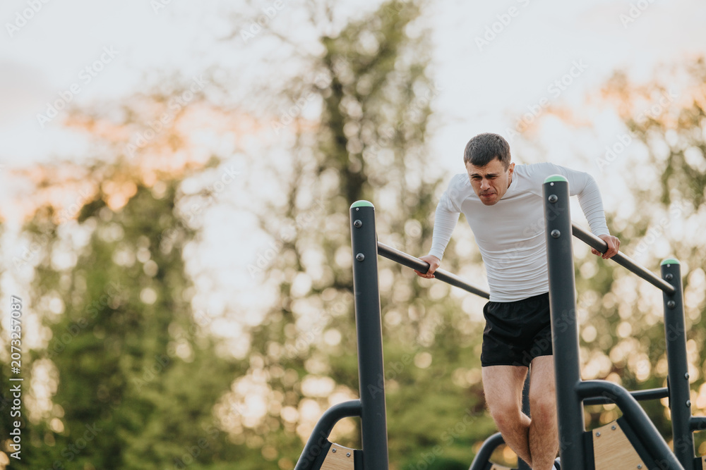 Fototapeta premium A man performs a dip exercise on parallel bars while training outdoors. He works on strength and balance during a park workout at sunset.