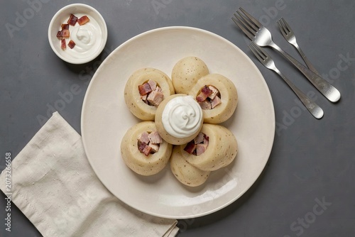 Cepelinai on white plate, topped with creamy sour cream. Diced bacon in small bowl, two silver forks beside. Concept of potato dumplings stuffed with pork bacon served elegantly.
