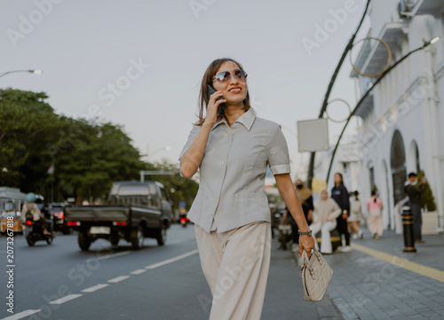 Fashionable style adult Asian Businesswoman commuting in sunglasses and casual button down shirt talking by phone call while walking in city at sunsets.