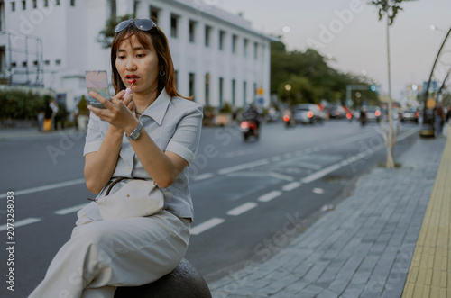 Fashionable adult Asian woman in black shirt casual clothes with batik bottom sits on a bench sidewalk applying lipgloss while looking in the mirror.