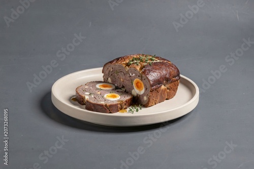 Drob de Miel on white plate with golden-brown crust, soft yolk oozing out. Concept of lamb offal loaf with herbs eggs presented elegantly against grey background.