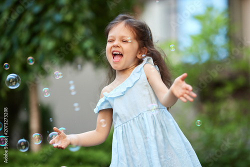 Cheerful young girl laughing, having fun playing with soap bubbles in a park, enjoying carefree summer childhood