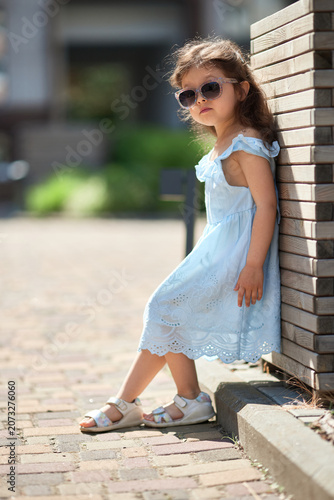 little child girl in a light blue sundress and sandals poses on a sunny urban sidewalk, playful and carefree on a warm summer day