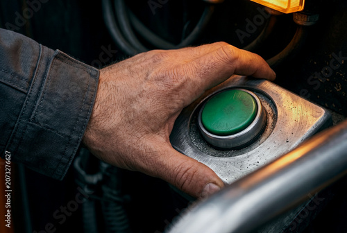 Macro eye-level focus on a muscular hand pressing a green mechanical start button. localized warm amber illumination highlights rugged skin relief, realistic hydration, and metal micro topography.
