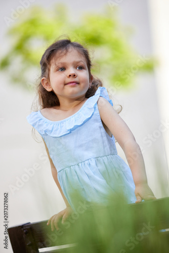 Little girl in a blue dress with a curious expression, looking away in a sunny outdoor setting, embodying childhood innocence
