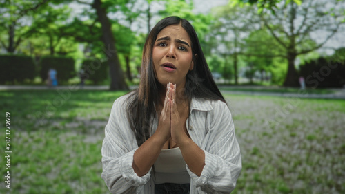 Obraz na plátně Woman showing hands in pleading fingertip and clasping gesture on street near park trees with worried face and open palms; desperation vulnerability