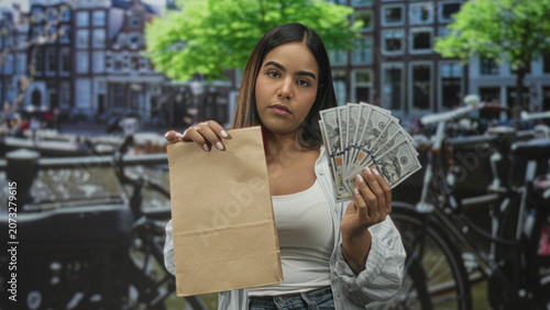 Foto Young woman holds brown paper bag and fans us dollars near bicycles and canal buildings on street; assertive transaction