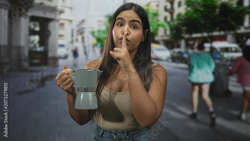 Photography Young woman holding coffeemaker while shushing with finger to lips and briefly raising an index finger on a busy street; playful secrecy