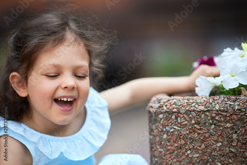 Smiling little girl in a blue dress playing with white and purple flowers in an outdoor planter, enjoying childhood joy and summer