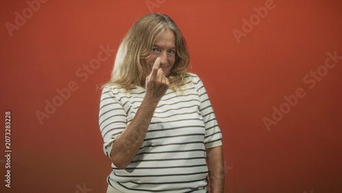Photos Senior woman blond smiling, wearing striped shirt, holding up middle finger with bare hand and relaxed posture against plain red studio wall; defiance
