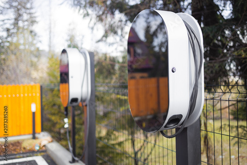 close-up shot of a modern EV home charging station wallbox with a wrapped cable, mounted on a pole outdoors next to a private house backyard.