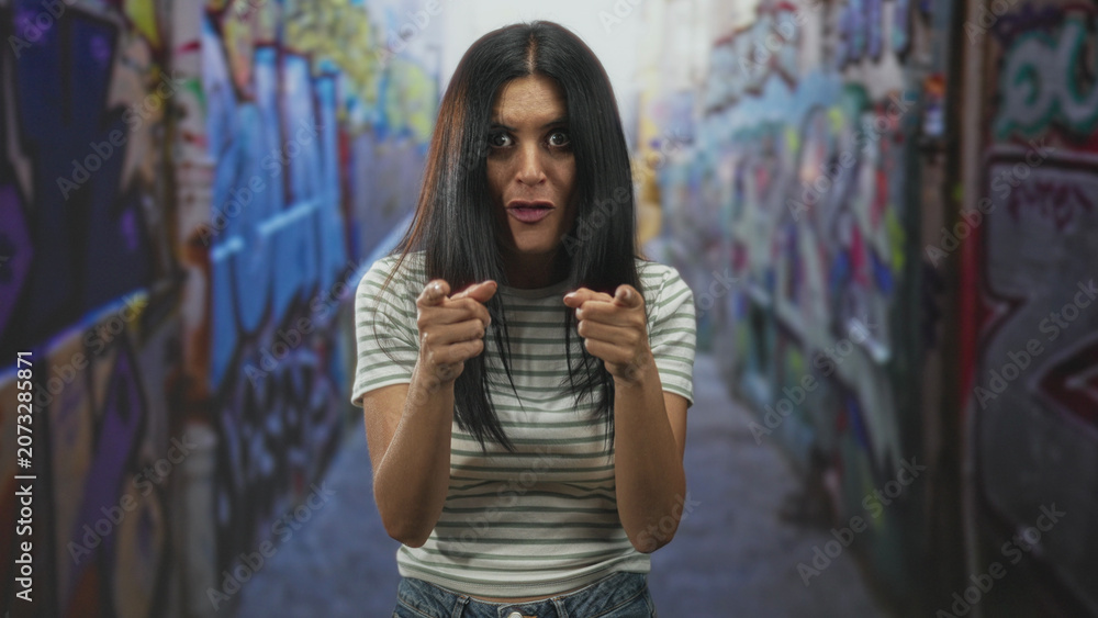 Naklejka premium Woman pointing both fingers with bare hands in graffiti street alley, direct gaze and open mouth; playful confidence.