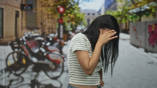 Canvastavla Young hispanic woman covering face with hand, palm to forehead on a street with parked bicycles and graffiti by buildings; shame vulnerability