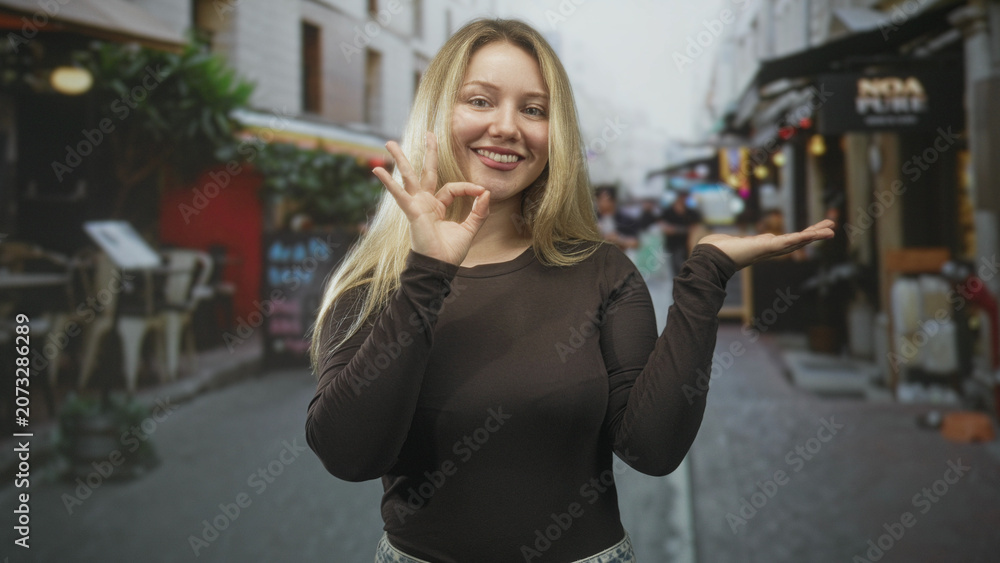 Fototapeta premium Woman making ok sign with hand and open palm while smiling in front of a street cafe and shop, presenting with approachable pose; confidence friendliness.