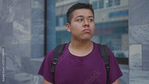 Fotografie Hispanic man with backpack standing by a stone building, face pouting with pursed lips and purple shirt visible; skepticism