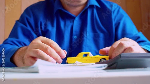 Car insurance agent playing with small yellow toy car at desk close up. Focus on hands and vehicle, shallow DOF, natural light. Bored businessman playing with miniature auto, procrastinating at work.