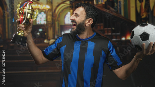 Photography Man holds trophy in left hand and soccer ball in right, mouth open in cheer, beard visible, standing in studio with ornate arches and stained glass backdrop; triumph