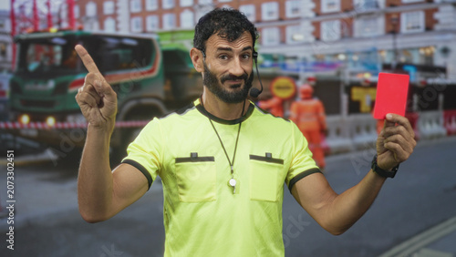 Fotografie Man referee holding red card and pointing index finger on street near construction lane with truck and workers; firm authority
