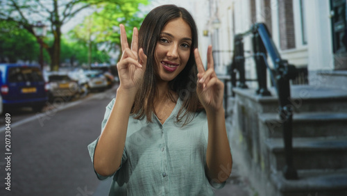 Fotografie Hispanic woman brunette smiling and showing peace sign with both hands on a city street stoop near parked cars; playful confidence