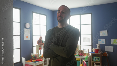 Canvastavla Man arms crossed smiling among toys and shelves in a bright kindergarten playroom building; calm reassurance