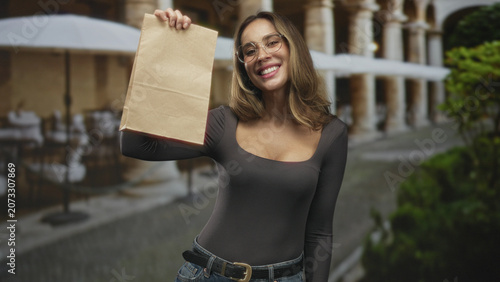 Foto Woman holds paper bag aloft and smiles showing cleavage in a street cafe with arches and umbrellas; joyful delivery