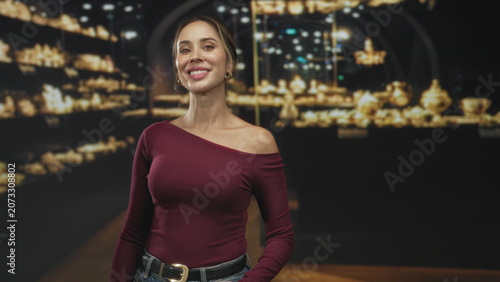 Принти на полотні Woman with exposed shoulder and hands in pockets smiling, wearing burgundy top before museum display in a building; quiet confidence