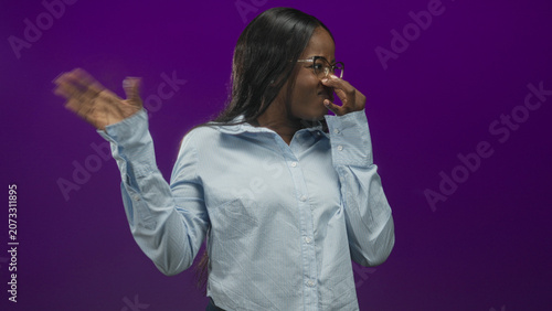 Fotografie Woman wearing glasses and light blue shirt pinching nose and waving hand in purple studio reacting to a foul smell; disgust avoidance