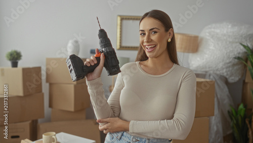 Billede på lærred Woman holding cordless drill aloft among stacked moving boxes in a building, unpacking and arranging belongings; confidence