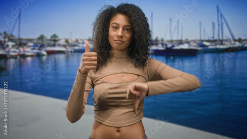 Obraz na plátne Woman giving thumbsup with visible navel on a street near marina boats and dock; indifference self expression